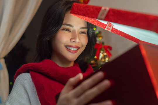 Cheerful Asian woman sitting on a sofa, excitedly opening a large red gift box at home with Christmas decorations in the background, holiday celebration.