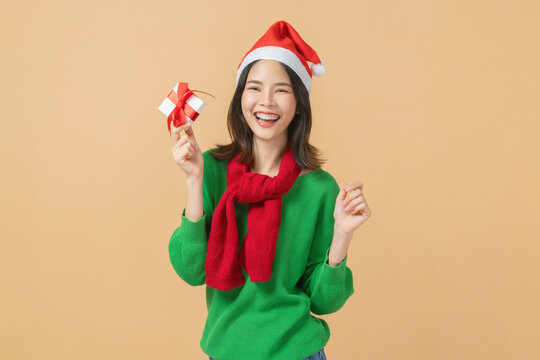 Cheerful Asian woman wearing a Santa hat, green sweater, and red scarf and holding gift box, Christmas celebration, festive holiday spirit concept.