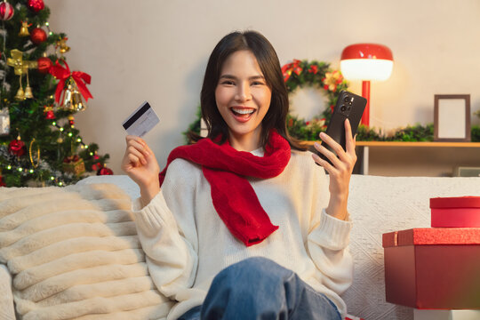 Cute Asian young woman wearing a white knitted sweater and red scarf, sitting on a sofa in the living room while using a smartphone and credit card for online shopping in Christmas day.