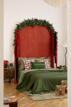 Cozy bedroom interior decorated for Christmas with a red accent wall, pine branch garland, festive pillows, green bedding, and warm candlelight glow