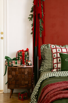Cozy festive bedroom interior decorated for winter holidays with red accent wall gift boxes on wooden nightstand and pine garland hanging above bed frame now
