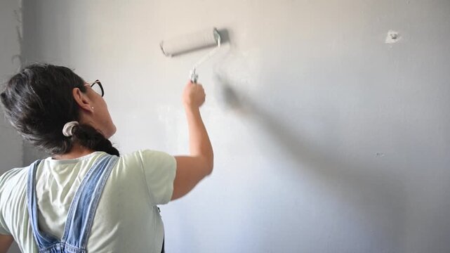 Woman applying gray paint to an interior wall using a paint roller during a DIY home renovation project. Home improvement, remodeling and interior decorating concept.