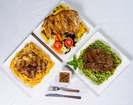 Peruvian food variety set top view with pesto green noodles huancaina pasta and native yellow fries served with grilled beef steak chicken chop hawaiian style set on white wooden background overhead f