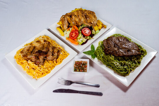 Peruvian food variety set top view with pesto green noodles huancaina pasta and native yellow fries served with grilled beef steak chicken chop hawaiian style set on white wooden background overhead f
