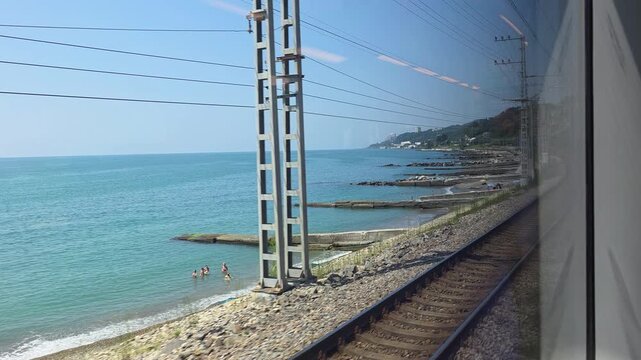 Train window framing the black sea shoreline, with swimmers, beach umbrellas and a pebble beach beside coastal railway tracks during a summer journey through sochi, russia