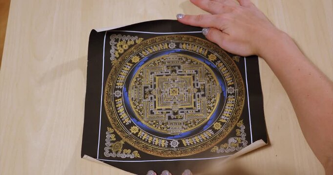Traditional Nepalese mandala scroll is unrolled upward by a hand in an overhead view exposing golden squares and blue rings with Buddhist symbols on black before hands hold it open
