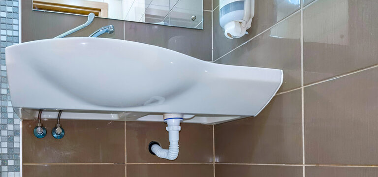 White bathroom sink with faucet and exposed plumbing against tiled wall. Modern washbasin in a public restroom. Bathroom fixture and interior design. Sanitation and personal hygiene
