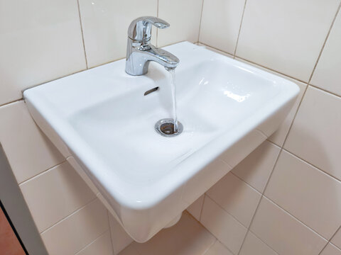 Water running from chrome faucet into white ceramic bathroom sink. Handwashing and personal hygiene. Household water use, plumbing fixture and bathroom sanitation