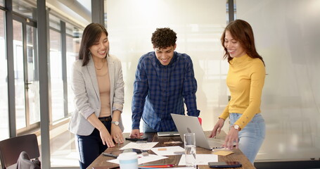 Diverse coworkers standing at wooden conference table reviewing documents and referencing laptop