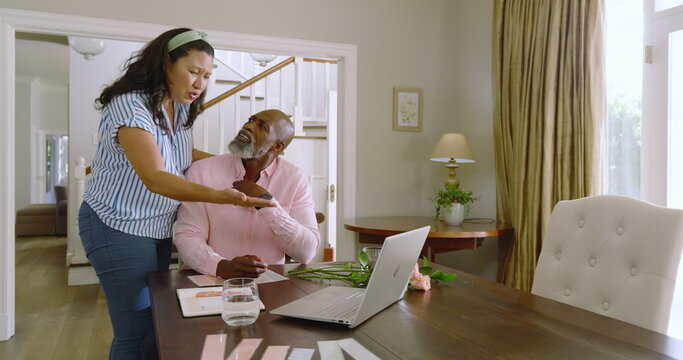 Senior diverse couple discussing plans at home, using laptop and documents on table