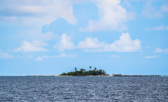 Small tropical island with green palm trees and white sand beach surrounded by blue ocean under cloudy sky on sunny day