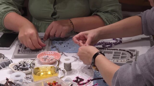 Two women crafting beaded jewelry together at table, close up view. Shared creative activity, handmade workshop. Friendship craft.