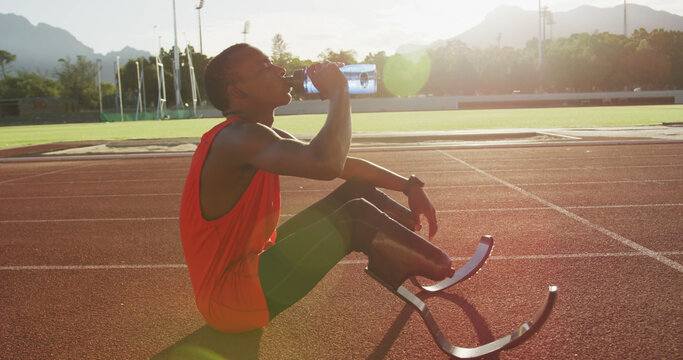 African american male athlete with prosthetic legs sitting and drinking water at empty stadium