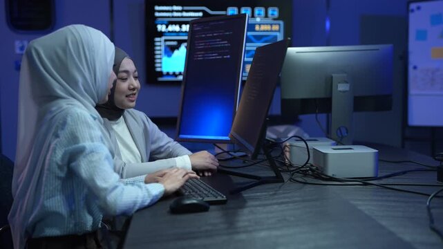 Two Muslim female software developers in hijab coding on monitors in dark office, Happy programmer team working on IT system, Modern technology, teamwork, cyber security and data development concepts