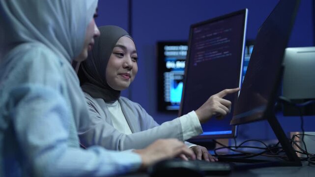 Two Muslim female software developers in hijab coding on monitors in dark office, Happy programmer team working on IT system, Modern technology, teamwork, cyber security and data development concepts