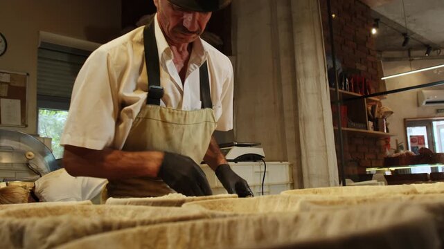 Elderly craftsman gives tender dough rounded form with scraper. Man places dough into sturdy rattan container designed for controlled proofing