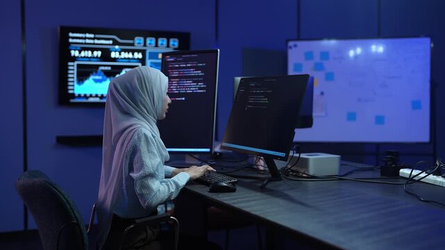 Muslim female software developer in hijab coding on multiple monitors in dark office, Happy programmer working on IT system and software development, Modern technology, cybersecurity and data concept