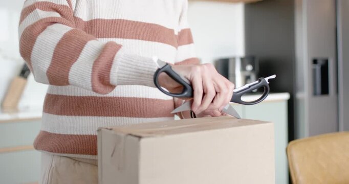 Person is lifting scissors, cutting box and using box cutter to reveal contents at kitchen counter