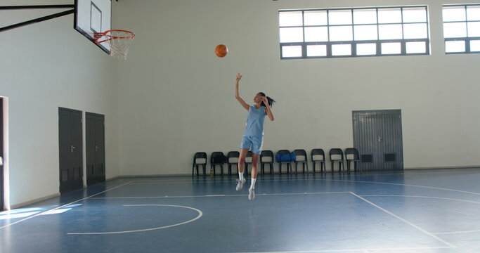Asian woman wearing light blue jersey jumping to shoot basketball at hoop in gymnasium with chairs