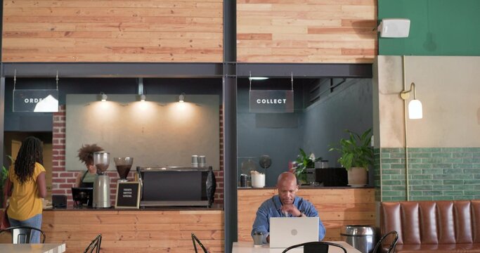 Typing male patron in blue button-up shirt at round table in cafe, laptop and takeaway cup