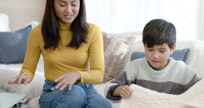 Asian mother and son practicing folding on sofa with gray basket after child lifting cream sweater