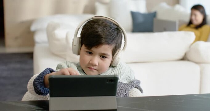 Asian child sitting at low table at home, leaning, tapping tablet in folio, headphones for learning