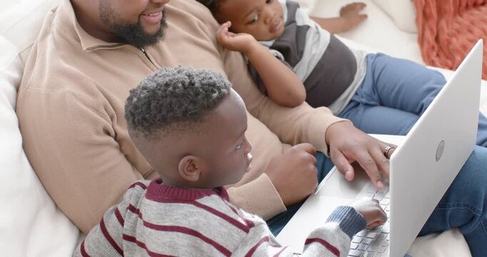 African American dad with two sons on couch teaching laptop after older child pressing keys