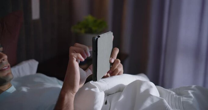 African American man lying in bed with duvet holding folio propping tablet tapping swiping browsing