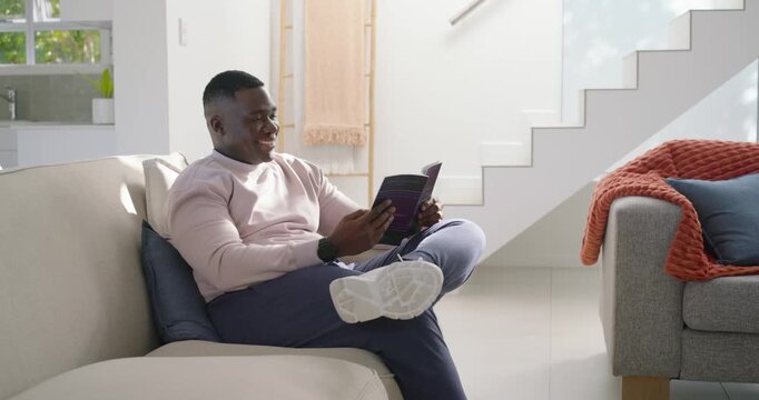 African man sitting on light colored sofa holding purple booklet and smiling while reading to relax