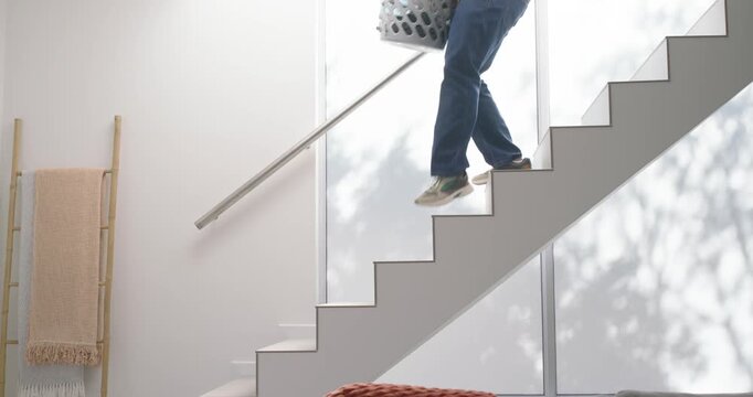 African man in yellow shirt carrying laundry basket down stairs into living area to fold clothes