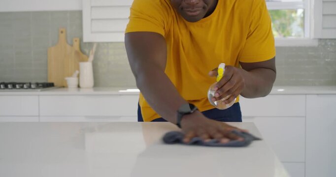 African man in mustard tee spraying bottle, pressing dark cloth and wiping kitchen island, cleaning