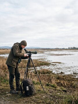 Birdwatcher using spotting scope on tripod at wetland with flock of birds. Wildlife observation and birding. Nature conservation and outdoor field research. Bird photography and ecotourism.