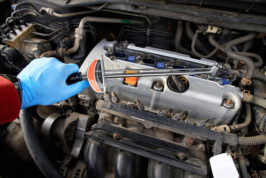 Hand in protective glove operating spark plug tool above silver valve cover. Mechanic using torque wrench tool on vehicle engine bay for spark plug service, tightening plugs with proper tension.