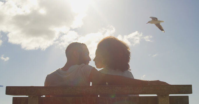 Romantic diverse couple sitting, embracing and kissing on sunny promenade, copy space