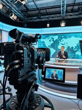 Professional video camera filming news anchor at broadcast studio desk with digital world map. Television news production and live reporting. Journalism, media and global current affairs.