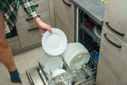 A man takes a white ceramic plate from a built-in dishwasher loaded with clean dishes and cookware