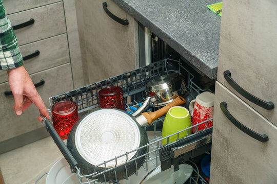 A man arranges red drinking glasses, colorful mugs, a frying pan, and cookware inside the open rack of a built-in dishwasher