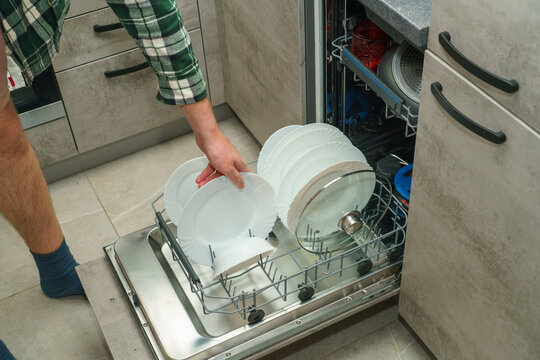A man takes a white ceramic plate from a built-in dishwasher loaded with clean dishes and cookware