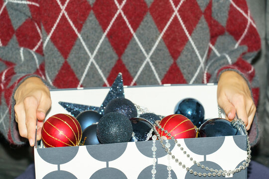 A woman holds a decorative storage box filled with red and navy blue Christmas baubles, glittering star ornaments, and silver bead garlands