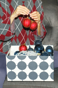 Close-up of a woman holding two red Christmas baubles decorated with elegant golden stripes above a box of holiday ornaments