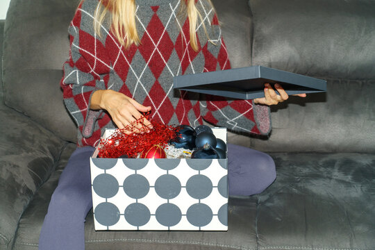 A woman sitting on a gray sofa opens a storage box filled with Christmas baubles and red decorative garland. The seasonal scene captures the preparation of holiday decorations at home