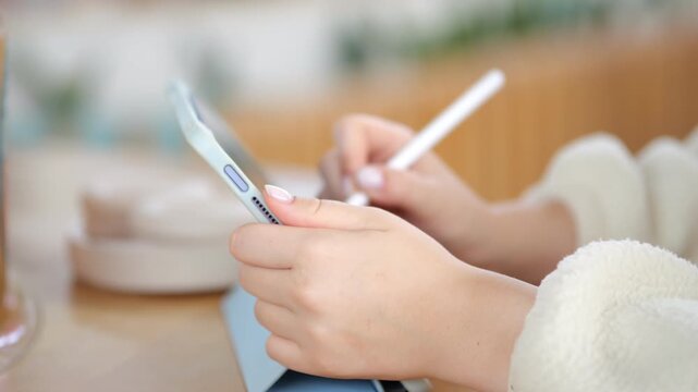 Close-up hands use stylus on tablet. Person wears soft white sweater. Background shows wooden table and blurred cafe setting. Focus remains on hands and device interaction