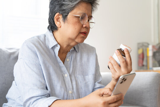 Anxious Asian senior woman holding medicine bottle and smartphone on sofa at home, checking drug information online, reading prescription or using telehealth application