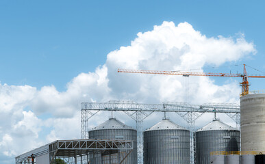 Agricultural grain silos and animal feed factory under construction with tower crane beneath a bright blue sky, building industrial storage infrastructure for commercial livestock feed production. © Fahroni