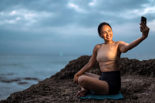 Asian woman having a video call on smartphone sitting on a cliff at sunset