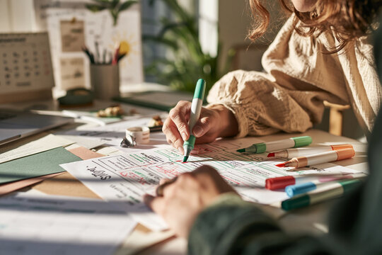 Two individuals collaborate at a sunlit desk focusing on a paper project draft One person uses a teal marker to annotate a detailed diagram while papers markers and supplies surround them