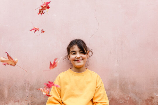 Smiling young girl in yellow sweatshirt with falling autumn leaves against pink wall background