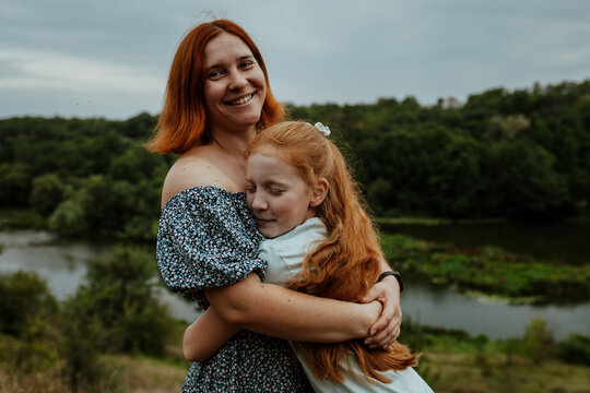 Happy mother hugging redhead daughter on hill above river