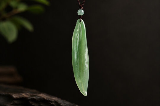 A slender green jade pendant shaped like a silique on a dark background.