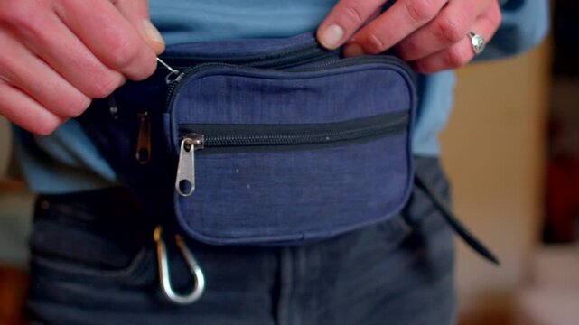 Close-up of person fastening navy fabric waist pouch at hip during casual indoor moment. Hands with ring and watch adjust zippers and press pouch flat against